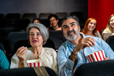 Senior couple watching a movie and eating popcorn, enjoying a night out at the cinema with other peopleの写真素材