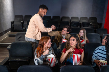Young people enjoying a movie night out at the cinema, greeting each other and eating popcornの写真素材