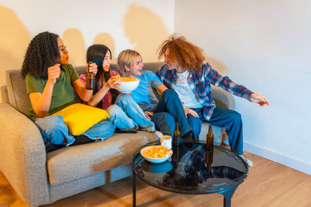 Four young friends enjoying their free time watching television, eating snacks and drinking beer, sitting on a comfortable sofaの写真素材