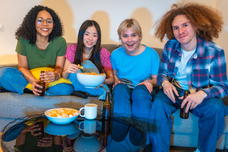 Four friends relaxing on a couch, sharing snacks and drinks while enjoying a movie together in a cozy living room settingの写真素材