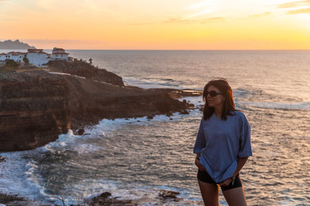 Young woman standing on cliff overlooking ocean waves crashing against rocky coastline at sunset in...