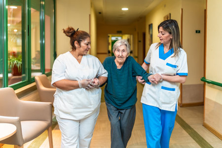 Two nurses assisting an elderly woman walking down the corridor of a nursing home, providing support and careの写真素材