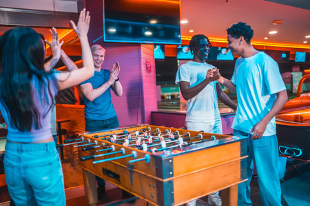 Multi ethnic friends enjoying a game of foosball, celebrating their victory with high fives and...