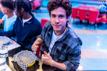 Young man gesturing thumbs up while sitting with friends at a bowling alley restaurant, enjoying a...