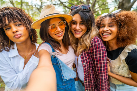 Four young women smiling and capturing a joyful selfie in a park, celebrating their friendship and enjoying a carefree moment togetherの写真素材