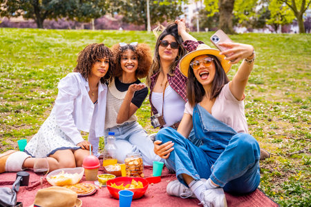 Four cheerful young women enjoying a sunny picnic in the park, capturing joyful moments together...