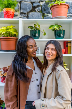 Two young businesswomen smiling together in front of a shelf adorned with vibrant plants, showcasing their friendship and professional partnershipの写真素材