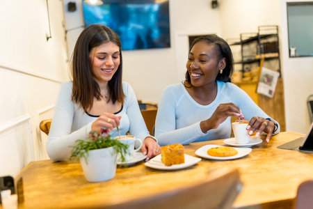Two young women enjoying a coffee break, chatting and laughing together in a vibrant, modern cafe filled with natural lightの写真素材
