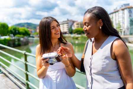Two female friends looking at a vintage camera, standing next to a river in a sunny city, enjoying their vacation togetherの写真素材