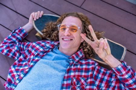 Smiling young man with curly hair relaxing on a skateboard, flashing a peace sign while lying on a...