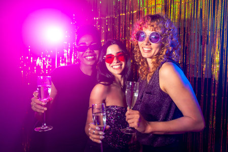 Three young women holding champagne glasses and smiling, enjoying a disco party with pink and blue lightsの写真素材