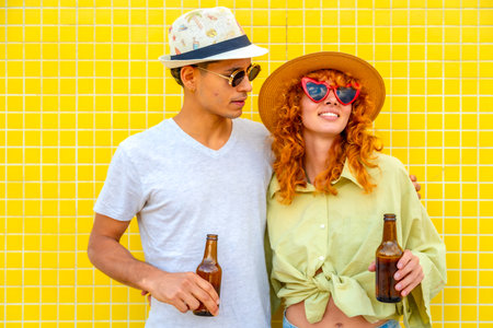 Young couple wearing hats and sunglasses, holding beer bottles and smiling joyfully against a vibrant yellow tiled wall, radiating happinessの写真素材