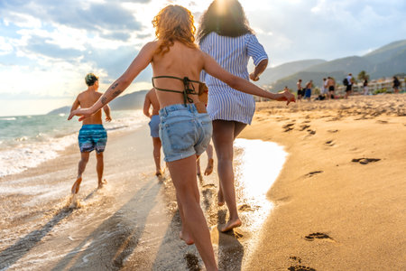 Group of friends running joyfully along the beach at sunset, savoring their summer vacation and the beauty of the ocean wavesの写真素材