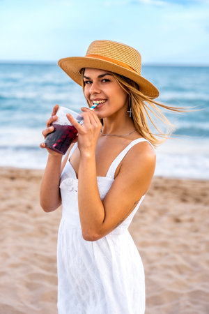 Young woman wearing straw hat and white dress drinking refreshing beverage on beach during summer vacationの写真素材