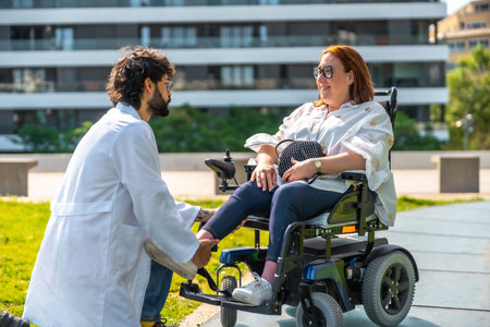 Doctor adjusting the footrest of an electric wheelchair for a smiling woman, enjoying a sunny day in the park surrounded by greeneryの写真素材