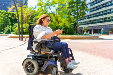 Happy woman enjoying a sunny day in the park, sitting in her wheelchair and browsing social media on...