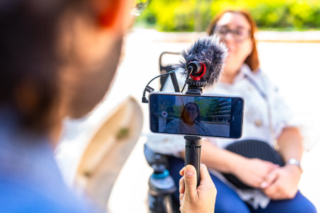 Videographer holding a smartphone on a tripod with external microphone, recording an interview with...