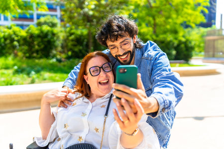 Cheerful couple taking a selfie in a park, the woman sitting on a wheelchair and the man embracing her, enjoying a sunny dayの写真素材