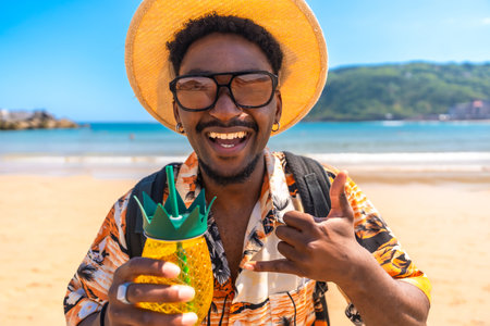 Young man enjoying summer vacation on a tropical beach, holding a pineapple shaped cocktail and gesturing the shaka signの写真素材