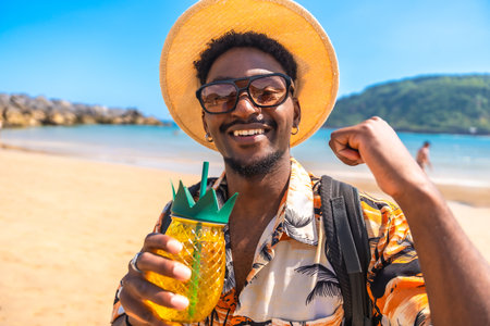 Young man smiling and raising fist while holding a pineapple shaped cocktail on a sunny beach, celebrating his summer vacationの写真素材