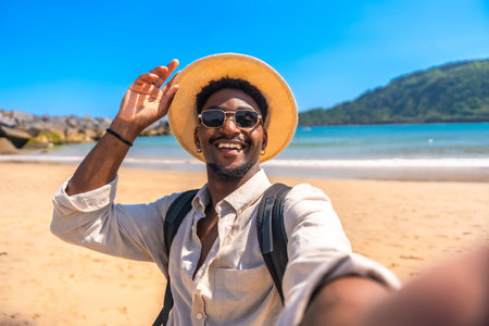 Young black man with backpack, sunglasses and straw hat taking a selfie on a tropical beach with turquoise waterの写真素材