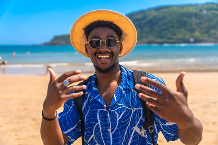 Young man wearing a hat and sunglasses, joyfully gesturing with his hands while enjoying the sunny tropical beach and ocean viewsの写真素材