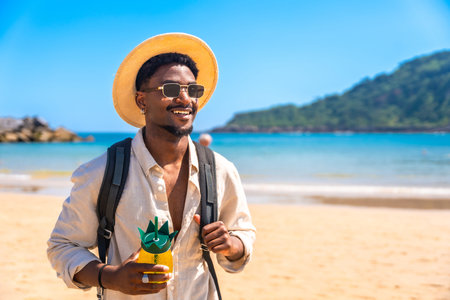 Young black man walking on a tropical beach, holding a pineapple shaped cocktail and wearing a...