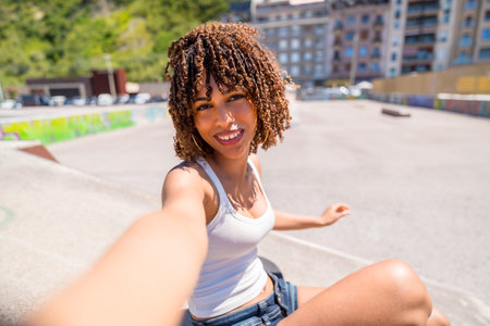 Smiling young woman with curly hair enjoying a sunny day at a skate park while taking a selfie portrait, capturing her vibrant urban lifestyleの写真素材
