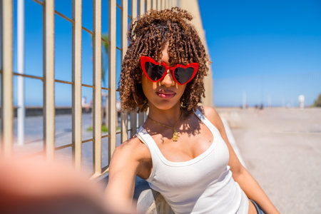 Fashionable young woman with curly hair taking a selfie by the beach, wearing trendy heart shaped sunglasses and a white topの写真素材