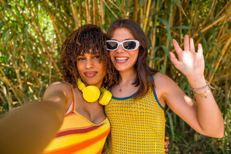 Two happy young women with headphones and sunglasses are taking a selfie and waving in a bamboo...
