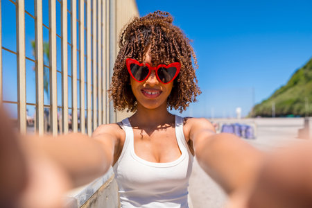 Smiling young woman with curly hair taking a selfie with heart shaped sunglasses on a sunny beach boardwalkの写真素材