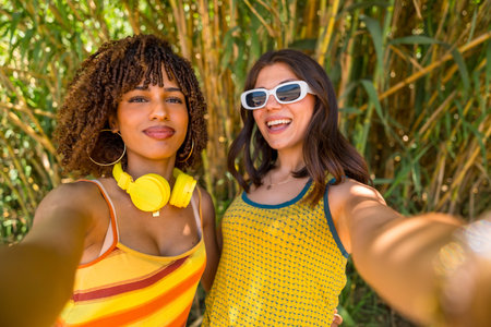 Two cheerful young women with colorful clothes and headphones taking a selfie in front of a bamboo forestの写真素材