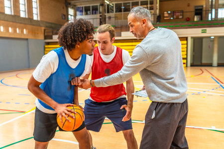 Basketball coach holding clipboard explaining game strategy to two players practicing during training in a gymの写真素材