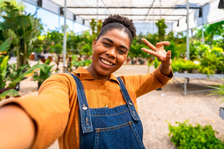 Happy young woman gardener taking a selfie and making a victory sign in a greenhouse full of plantsの写真素材