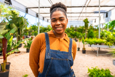 Portrait of a young African American woman smiling while working in a greenhouse, surrounded by vibrant plants and flowersの写真素材
