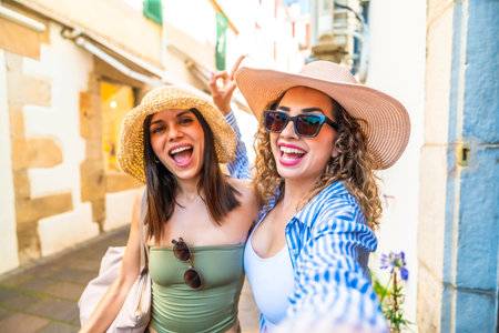 Two cheerful female tourists wearing summer hats and sunglasses are taking a selfie with a cheerful expression while walking down a city street during their vacationの写真素材