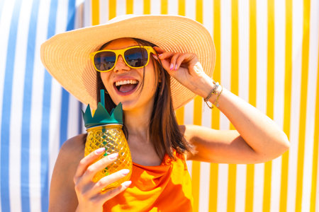 Young woman with sunglasses and a straw hat, enjoying a tropical pineapple juice on a colorful...