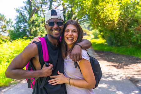 Interracial couple wearing backpacks and embracing while enjoying a hike on a sunny nature trailの写真素材