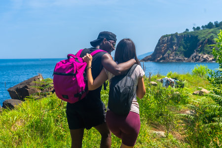 Interracial couple with backpacks walking on a path by the sea, embracing and enjoying the scenic...