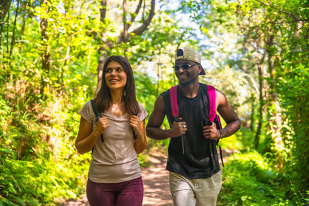 Interracial couple wearing backpacks walking in a forest trail, enjoying the beautiful nature...