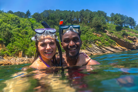 Interracial couple snorkeling in the clear blue sea, surrounded by a stunning green cliff, enjoying a joyful summer adventure togetherの写真素材