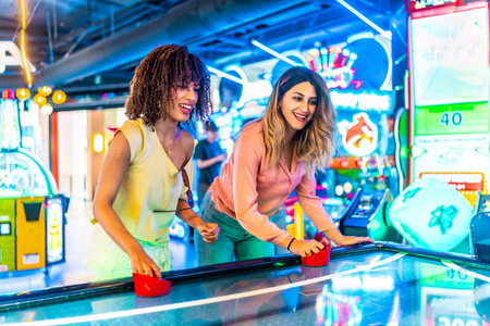 Two young women enjoying a friendly game of air hockey in a vibrant arcade, illuminated by colorful neon lightsの写真素材