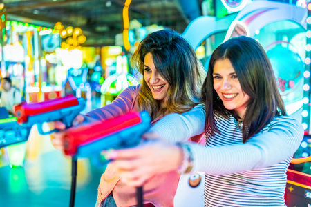 Two young women enjoying leisure time, playfully engaging with toy guns in a vibrant amusement arcade filled with colorful lights and laughterの写真素材