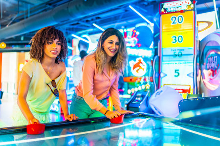 Two young women enjoying a friendly game of air hockey in a vibrant arcade, illuminated by colorful neon lightsの写真素材