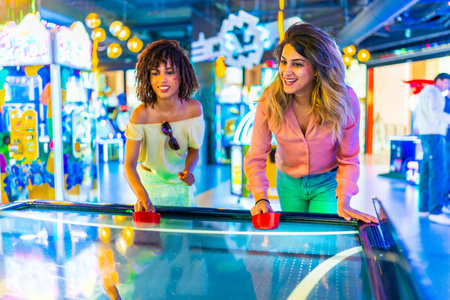 Two female friends enjoying a lively game of air hockey in a vibrant amusement arcade filled with colorful lights and excitementの写真素材