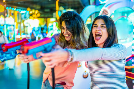 Two women laughing and enjoying their free time while playing arcade games with colorful toy guns, surrounded by vibrant lights and excitementの写真素材