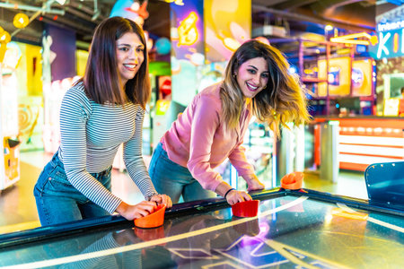 Two female friends enjoying their free time playing air hockey in a colorful and modern amusement arcadeの写真素材