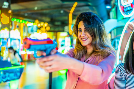 Cheerful young woman holding a toy gun, playing at an arcade game machine, enjoying her free time with friendsの写真素材