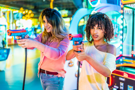 Two women laughing and enjoying their free time while playing colorful arcade shooting games, immersed in fun and friendly competitionの写真素材