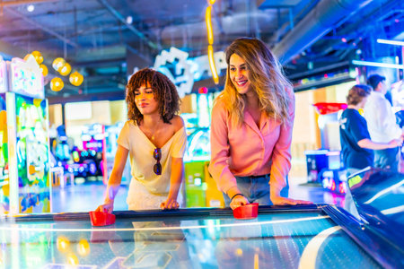 Two female friends concentrating while playing air hockey in a vibrant arcade, enjoying their leisure timeの写真素材